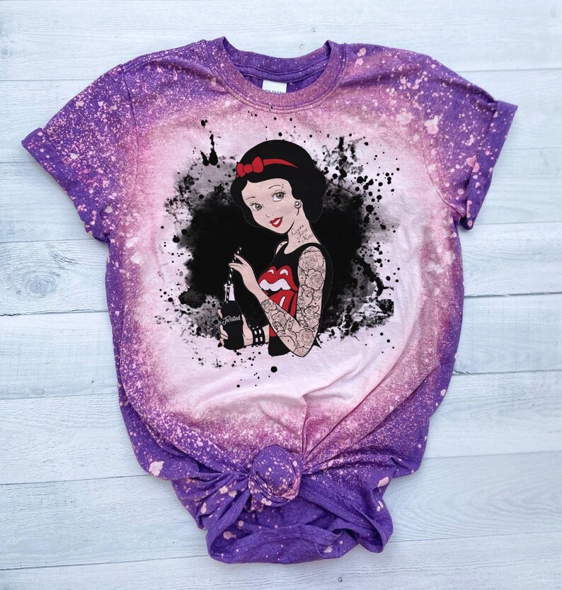 May include: Purple and pink tie-dye t-shirt with a black and white graphic of Snow White with tattoos and holding a bottle of soda. The graphic has a splatter effect.