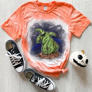 Op de afbeelding: Oranje gebleekt T-shirt met een groen en zwart cartoonfiguur op de voorkant. Het figuur danst op een stekelig platform. Het shirt is onderaan geknoopt. Er zijn twee zwarte Converse schoenen en een witte pompoen met een zwart gezicht op de tafel.