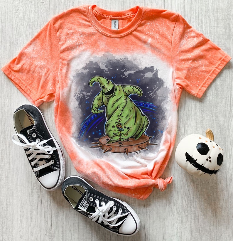 Op de afbeelding: Oranje gebleekt T-shirt met een groen en zwart cartoonfiguur op de voorkant. Het figuur danst op een stekelig platform. Het shirt is onderaan geknoopt. Er zijn twee zwarte Converse schoenen en een witte pompoen met een zwart gezicht op de tafel.