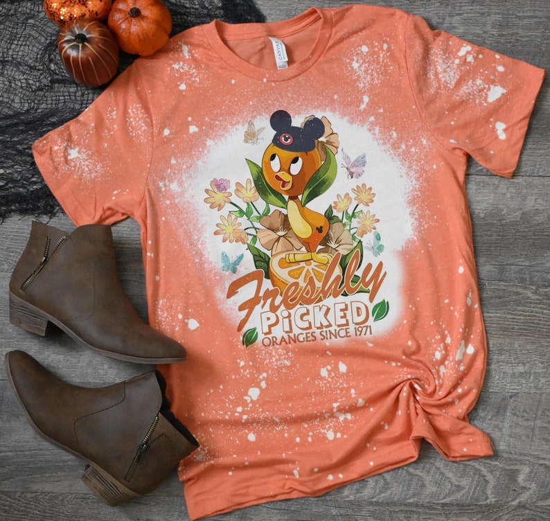 May include: Orange t-shirt with a bleach-splattered design featuring a cartoon orange with Mickey Mouse ears and the text "Freshly Picked Oranges Since 1971".