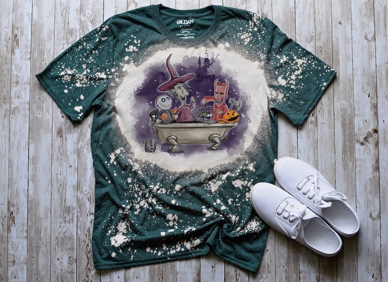 K&ouml;nnte beinhalten: Ein gr&uuml;nes T-Shirt mit einem bleichmittelgesprenkelten Design, das Figuren aus dem Film Nightmare Before Christmas zeigt. Die Figuren befinden sich in einer Badewanne mit K&uuml;rbissen und einem K&auml;fig. Das Shirt hat ein Gildan-Etikett.