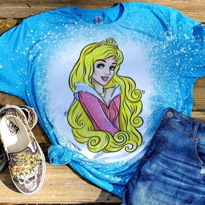 May include: A blue t-shirt with a bleach-splattered design featuring a cartoon image of Sleeping Beauty. The image is in yellow, pink, and white.