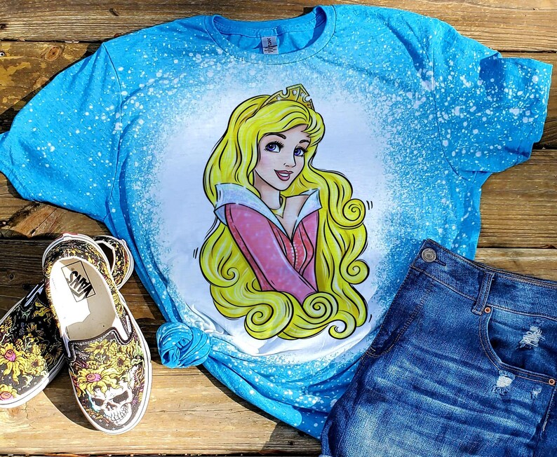 May include: A blue t-shirt with a bleach-splattered design featuring a cartoon image of Sleeping Beauty. The image is in yellow, pink, and white.