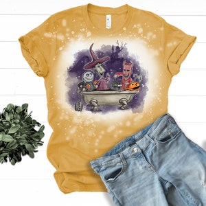 K&ouml;nnte beinhalten: Ein gelbes T-Shirt mit einem Design aus Bleichmittelspritzern, das ein Cartoon-Bild von Figuren aus dem Film Nightmare Before Christmas zeigt. Die Figuren befinden sich in einer Badewanne mit K&uuml;rbissen und anderen Halloween-Dekorationen.