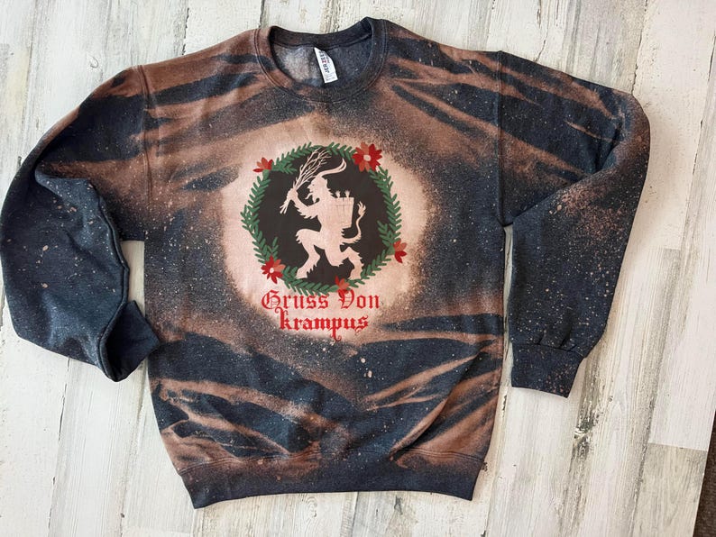 May include: Black and brown tie-dye sweatshirt with a white silhouette of a Krampus figure inside a wreath with red berries and the text "Gruss Von Krampus".