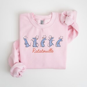 May include: A pale pink sweatshirt with a cartoon illustration of five blue rats in chef hats. The word "Ratatouille" is written in red script beneath the rats. The sweatshirt has a crew neck and long sleeves.