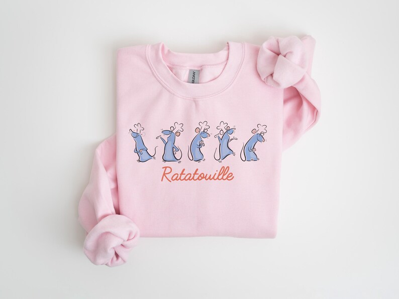 May include: A pale pink sweatshirt with a cartoon illustration of five blue rats in chef hats. The word "Ratatouille" is written in red script beneath the rats. The sweatshirt has a crew neck and long sleeves.