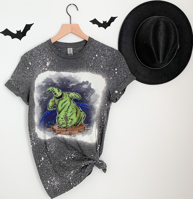 Op de afbeelding: Een grijs T-shirt met een bleekmiddel-gespatte ontwerp met een groen personage uit The Nightmare Before Christmas. Het personage staat op een platform met spikes.
