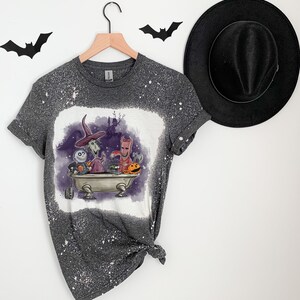 K&ouml;nnte beinhalten: Ein graues T-Shirt mit einem bleichmittelgesprenkelten Design, das Figuren aus dem Film Nightmare Before Christmas zeigt. Die Figuren befinden sich in einer Badewanne mit einem K&uuml;rbis und einem K&auml;fig.