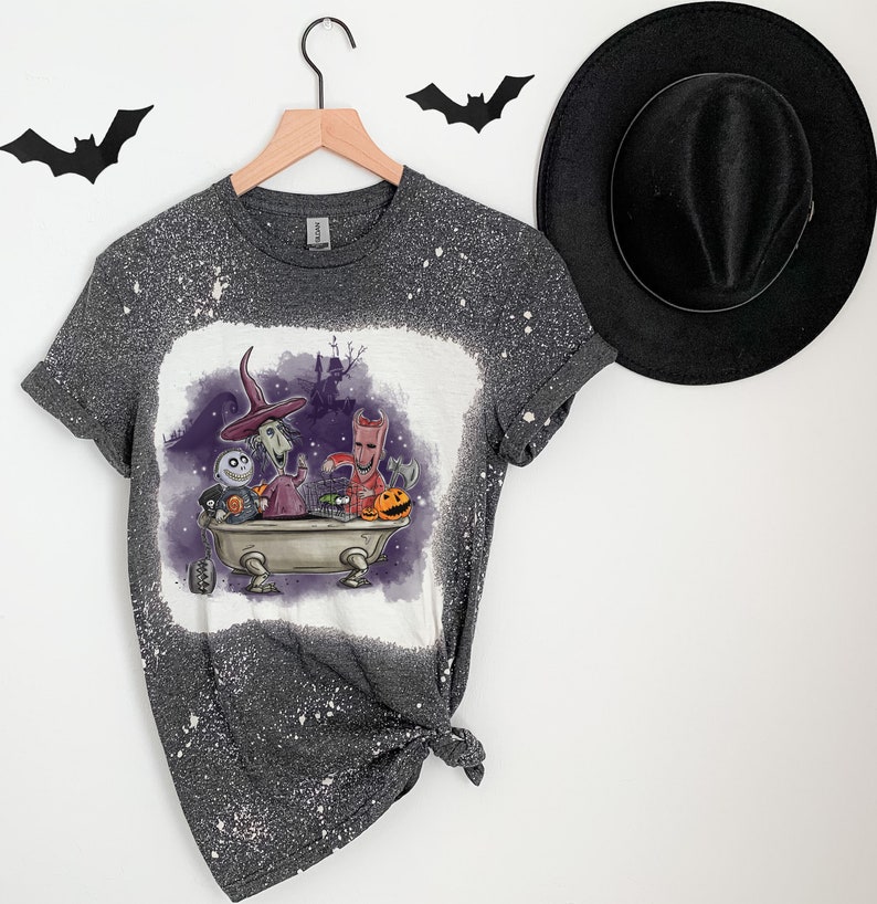 K&ouml;nnte beinhalten: Ein graues T-Shirt mit einem bleichmittelgesprenkelten Design, das Figuren aus dem Film Nightmare Before Christmas zeigt. Die Figuren befinden sich in einer Badewanne mit einem K&uuml;rbis und einem K&auml;fig.