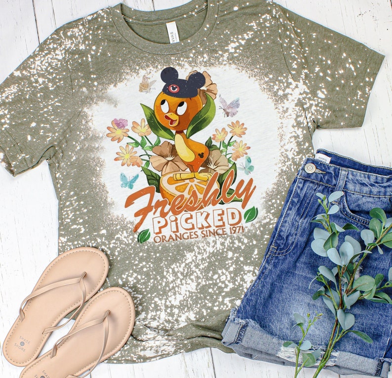 May include: A green t-shirt with a bleach-splattered design featuring a cartoon orange with Mickey Mouse ears and the text "Freshly Picked Oranges Since 1971".