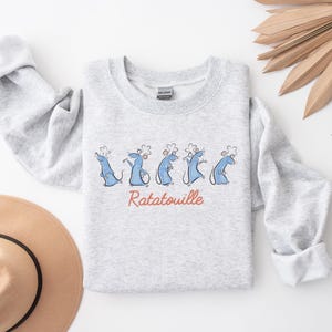 May include: A light grey sweatshirt with a cartoon design of blue rats wearing chef hats. The word "Ratatouille" is written in red script below the rats. The sweatshirt has long sleeves and a crew neck.