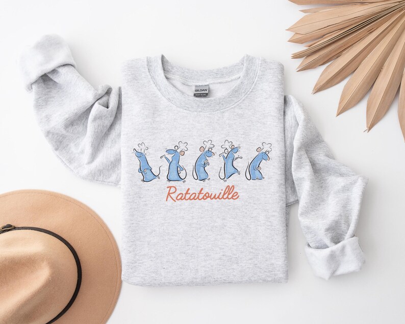 May include: A light grey sweatshirt with a cartoon design of blue rats wearing chef hats. The word "Ratatouille" is written in red script below the rats. The sweatshirt has long sleeves and a crew neck.
