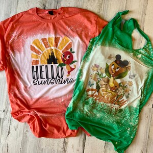 May include: Two bleached t-shirts with a Disney theme. The orange shirt has a sunset graphic with a castle and a bird, and the text "Hello Sunshine". The green shirt has a bird with a Mickey Mouse hat, flowers, and the text "Freshly Squeezed Oranges Since 1971".