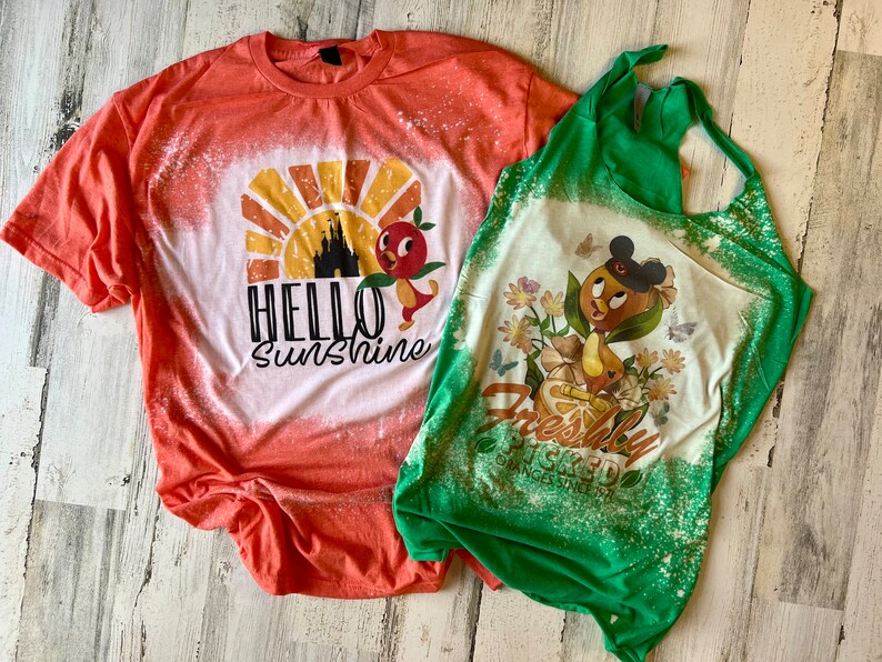 May include: Two bleached t-shirts with a Disney theme. The orange shirt has a sunset graphic with a castle and a bird, and the text "Hello Sunshine". The green shirt has a bird with a Mickey Mouse hat, flowers, and the text "Freshly Squeezed Oranges Since 1971".