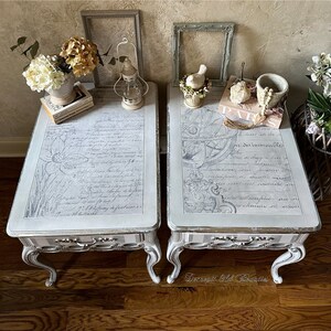 May include: Two matching side tables with a distressed white finish. The tabletops feature a floral and script design in gray. Each table has a drawer with a silver handle and curved legs. The tables are decorated with flowers and books.