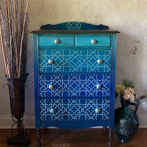 May include: A hand-painted chest of drawers in shades of blue, with a geometric pattern on the drawers. The chest has a dark brown top and decorative knobs. A decorative vase and rug are in the foreground.