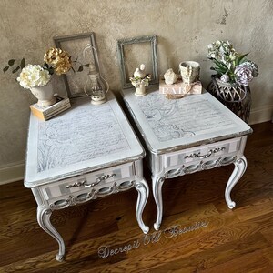 May include: Two white side tables with ornate silver trim and curved legs. The tabletops feature a floral and script design. Each table has a single drawer with silver hardware. The tables are decorated with flowers and framed artwork.