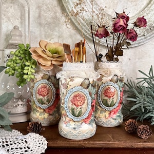 May include: Three decorative glass jars with floral designs and lace trim, used as a brush holder. The jars are filled with paintbrushes and are sitting on a wooden surface.