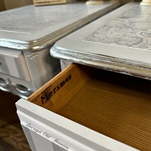 May include: Two silver and white painted bedside tables with drawers. The drawers are open on one table, revealing the word "Mersman" inside. The table tops have a decorative design. The bottom of the table has vertical stripes. The text "Decrepit Old Beauties" is on the drawer.