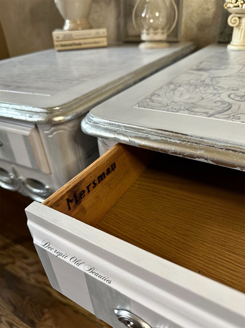 May include: Two silver and white painted bedside tables with drawers. The drawers are open on one table, revealing the word "Mersman" inside. The table tops have a decorative design. The bottom of the table has vertical stripes. The text "Decrepit Old Beauties" is on the drawer.