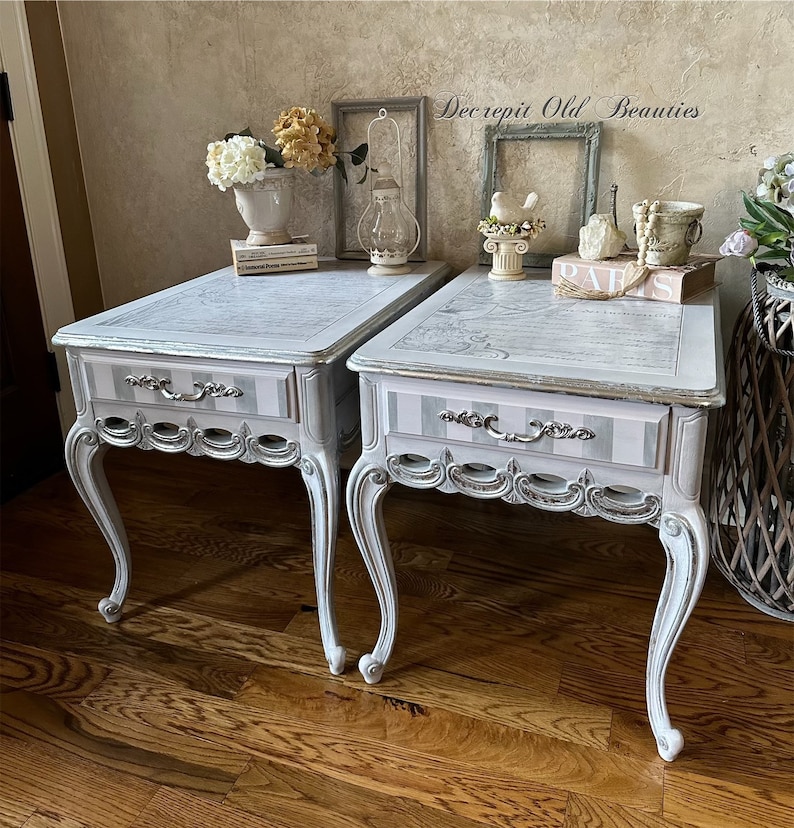 May include: Two matching side tables with a distressed, vintage aesthetic. The tables feature a light gray and white color scheme with a striped drawer front and ornate silver hardware. The tops have a map design. The legs are curved and painted white.