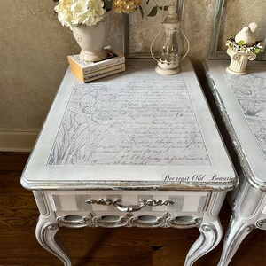 May include: Two matching antique side tables with a distressed gray finish. The tops feature a floral design and script. Each table has a drawer with silver hardware and curved legs. Decorative items include flowers, books, and a lantern.