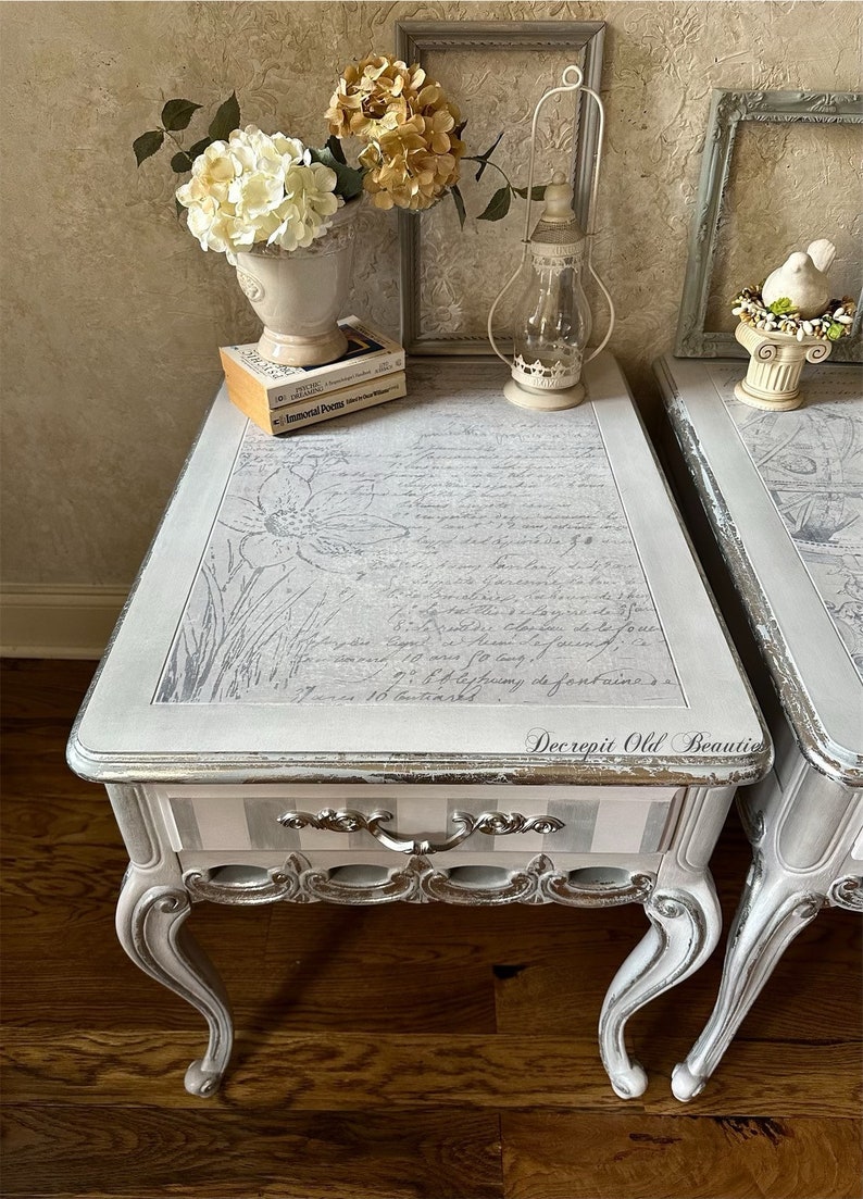 May include: Two matching antique side tables with a distressed gray finish. The tops feature a floral design and script. Each table has a drawer with silver hardware and curved legs. Decorative items include flowers, books, and a lantern.