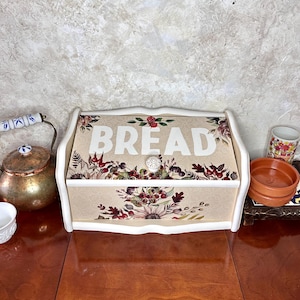 Puede incluir: Una caja de pan blanca con un diseño floral y la palabra "BREAD" en letras blancas en la parte delantera. La caja tiene un pomo blanco y está sobre una mesa de madera.