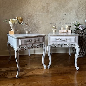 May include: Two matching white and gray side tables with ornate details. Each table features a drawer with a striped design and decorative hardware. The tables have curved legs and are placed on a hardwood floor.