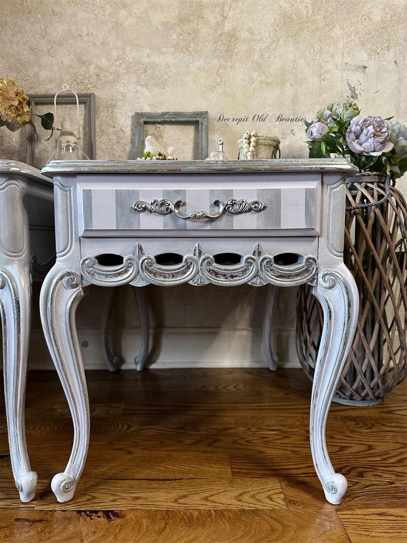 May include: A pair of antique-style bedside tables. Each table features a drawer with a silver handle and a striped design in gray and pink. The tables have ornate, curved legs and decorative trim, with the text "Decrepit Old Beauties" visible.