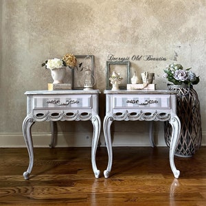 May include: Two matching antique bedside tables with white and gray striped tops and ornate, curved legs. Each table has a single drawer with a decorative handle. The tables are painted white with distressed detailing and are set against a neutral background.