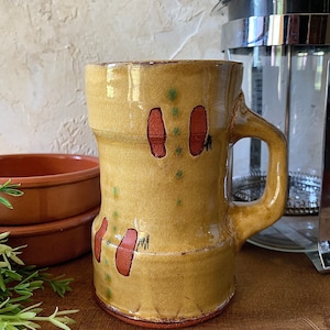 May include: A yellow ceramic mug with a brown handle and red footprints painted on the side. The mug has a textured surface and a slightly uneven rim.