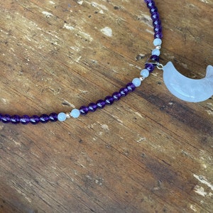 May include: A purple and white beaded necklace with a crescent moon charm. The charm is made of white quartz.