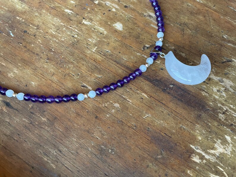 May include: A purple and white beaded necklace with a crescent moon charm. The charm is made of white quartz.