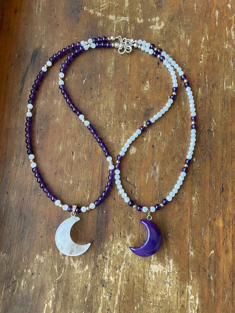 May include: Two crescent moon pendants on a layered necklace. The top layer is made of white beads and the bottom layer is made of purple beads.
