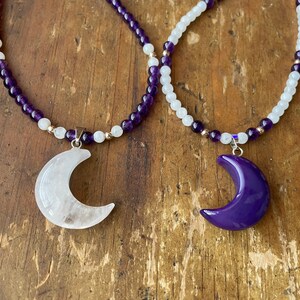 May include: Two necklaces with crescent moon pendants. One necklace is made of amethyst beads and has a white moon pendant. The other necklace is made of white beads and has a purple moon pendant.