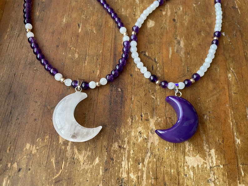 May include: Two necklaces with crescent moon pendants. One necklace is made of amethyst beads and has a white moon pendant. The other necklace is made of white beads and has a purple moon pendant.