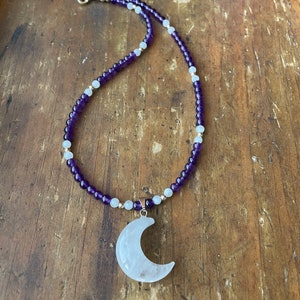 May include: A necklace made with amethyst and white beads with a crescent moon pendant made of clear quartz.
