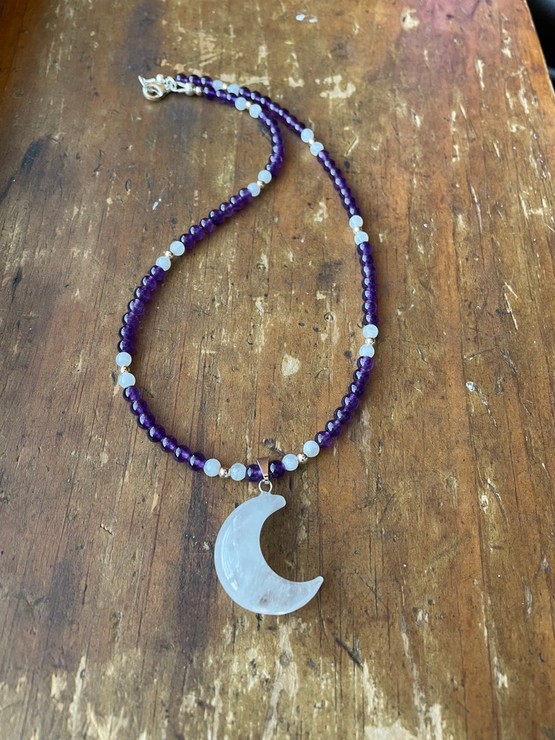 May include: A necklace made with amethyst and white beads with a crescent moon pendant made of clear quartz.