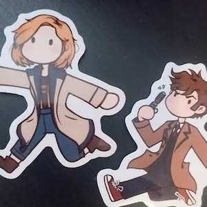 Peut inclure: Deux autocollants avec des illustrations de personnages de dessins animés. L'un représente un personnage en manteau beige et pantalon bleu. L'autre montre un personnage en manteau marron tenant un appareil. Les deux autocollants ont une bordure blanche.