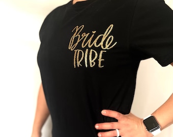Może przedstawiać: Czarny t-shirt ze złotym brokatowym napisem "Bride Tribe". Koszulka z krótkim rękawem ma okrągły dekolt. Zdjęcie przedstawia osobę noszącą koszulkę z ręką na biodrze.
