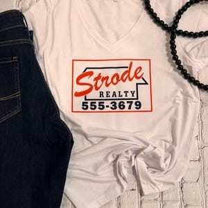 May include: White v-neck t-shirt with a red and black graphic that says "Strode Realty 555-3679".