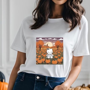 Puede incluir: Camiseta blanca con un gráfico de un gato de dibujos animados en un campo de calabazas. El gato lleva una camisa blanca y una corbata de lazo roja. El fondo es una escena de puesta de sol con colores naranja y amarillo.