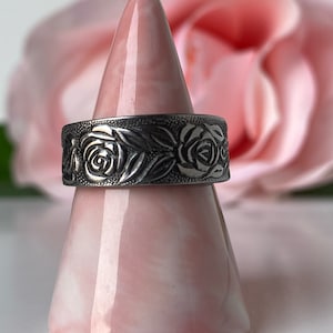 May include: A silver ring with a rose design. The ring is on a pink display stand.