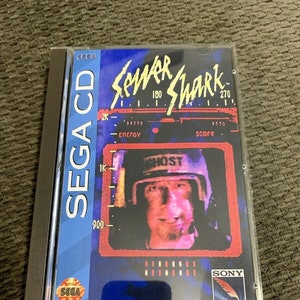 Puede incluir: Una caja de juego Sega CD para Sewer Shark, un videojuego para el sistema Sega CD. La caja es azul y presenta una captura de pantalla del juego con un personaje que lleva un casco. El texto "Sewer Shark" está en amarillo y el texto "Sega CD" está en blanco.