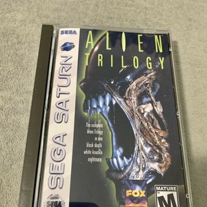 Puede incluir: Una caja de juego de Sega Saturn con una criatura alienígena negra y verde en la parte delantera. El título del juego es "Alien Trilogy" y la caja tiene una clasificación "Mature" de la ESRB.