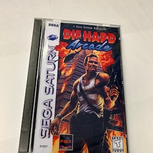 Könnte beinhalten: Ein Sega Saturn Videospiel-Etui mit einem rot-weiß-schwarzen Design. Das Spiel ist Die Hard Arcade, ein Sega Saturn-Exklusivtitel. Das Etui zeigt ein Bild von John McClane, der Hauptfigur aus der Die Hard-Filmreihe, der eine Waffe hält. Das Spiel ist von der ESRB mit T für Teenager bewertet.