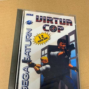 Puede incluir: Caja de juego Sega Saturn para Virtua Cop, un juego para 1-2 jugadores. La caja es de plástico transparente con una columna vertebral negra. La parte frontal de la caja presenta una imagen colorida de dos policías que sostienen armas de fuego, con un horizonte de la ciudad en el fondo. El texto "SEGA SATURN", "SEGA", "VIRTUA COP" y "1-2 PLAYER GAME" es visible en la caja. La clasificación ESRB "TEEN" también es visible en la caja.