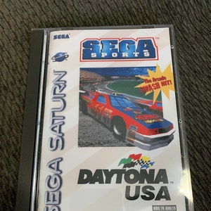 Puede incluir: Una caja de juego Sega Saturn para Daytona USA. La caja es blanca con un coche de carreras rojo, blanco y azul en la parte delantera. El juego está clasificado como "KA" para niños y adultos.